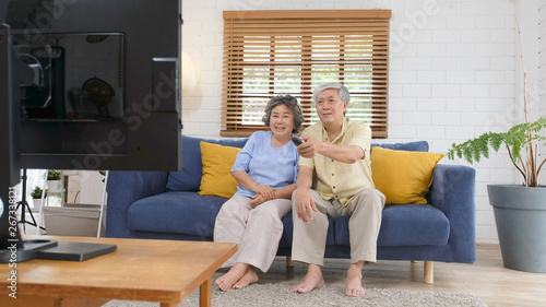 Senior asian couple holding remote control while watching television in home living room with happiness, old people retirement routine lifestyle