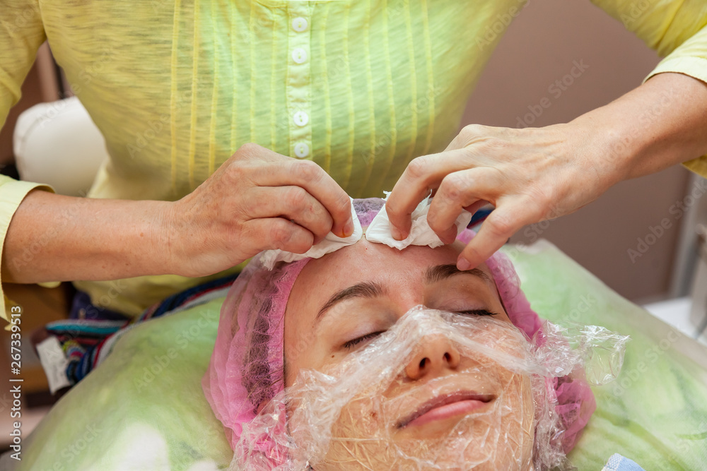 A young girl is lying on a couch during cosmetic procedures with a mask on the face above which beautician woman squeezes body fat and pimples with wipes. Cleansing and make up. A young girl is lying on a couch during cosmetic procedures with a mask on the face above which beautician woman squeezes body fat and pimples with wipes. Cleansing and make up.