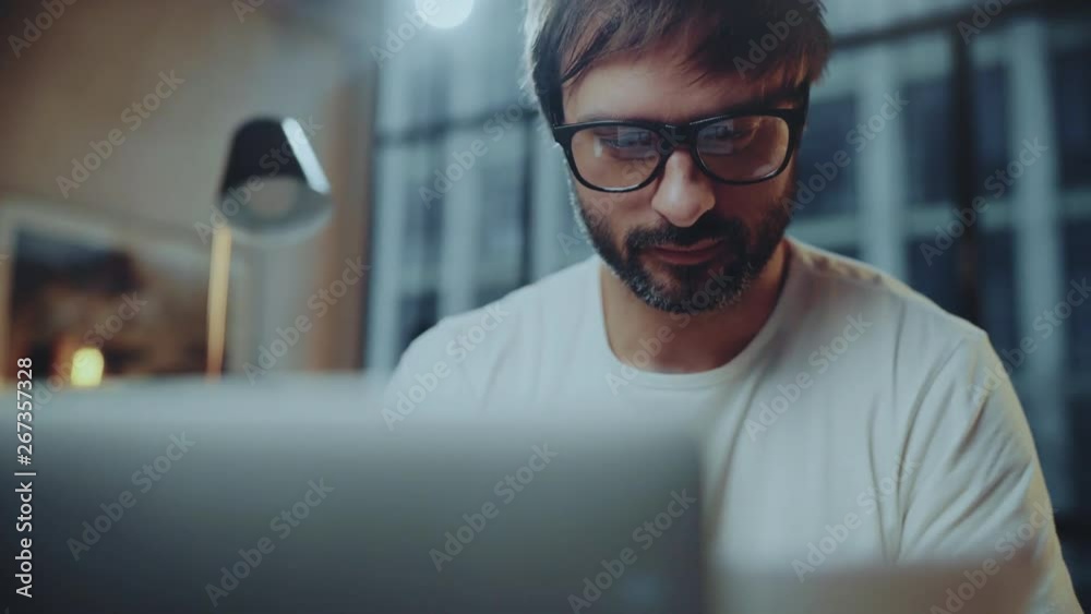 Young bearded professional coworker wearing eye glasses working at modern loft studio-office with laptop computer.Big panoranic windows on blurred background