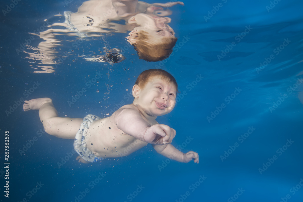 Funny face portrait of baby boy with tongue sticking out swimming and diving underwater with fun in the pool. Healthy family lifestyle and children water sports activity. Child development.