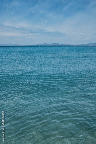 calm sea of turquoise color and some clouds in the blue sky, sailboats and mountains far away in the background