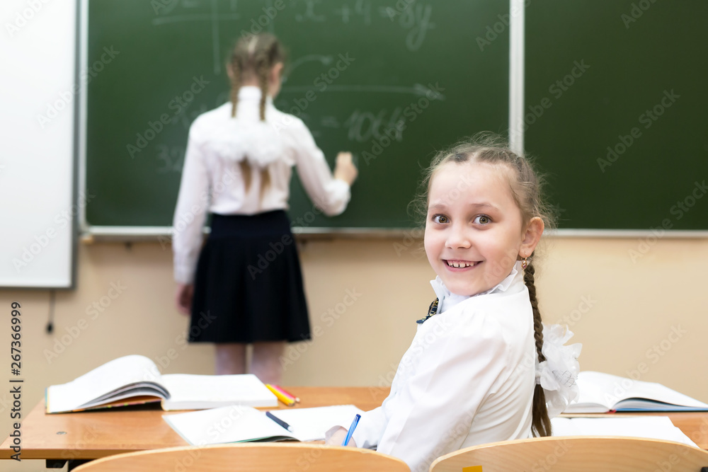 A schoolgirl girl looks into the camera while her friend, a classmate, answers the teacher at the blackboard A schoolgirl girl looks into the camera while her friend, a classmate, answers the teacher at the blackboard