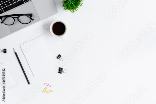 The office desk flat lay view with laptop, mouse, tree, pink and yellow paper clip, coffee cup, notebook, pencil, black glasses on white background.