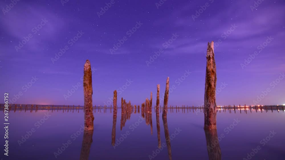 Timelapse of moving stars and clouds  in night sky and water reflection. The Milky Way galaxy rotating over the unusual wooden poles atb the water lake  in summer time