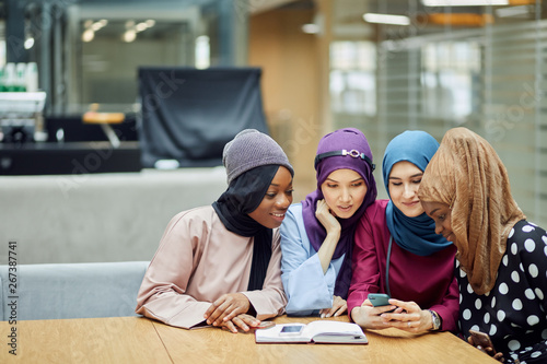 Beautiful young muslim multiethnic group of girls in long smart traditional dresses and hijabs looking at smart phone in hand of asian woman while studying at college lobby.