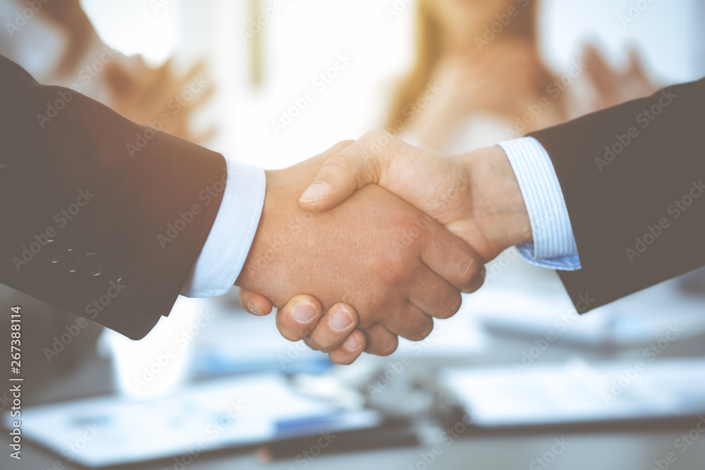 Business people shaking hands at meeting while theirs colleagues clapping and applauding. Group of unknown businessmen and women in modern white office. Success teamwork, partnership and handshake