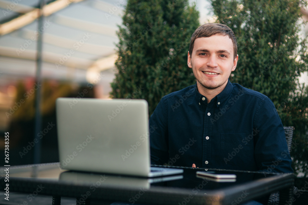 Businessman using laptop in cafe. Handsome man in dark shirn sit on the terrace. Background of green tree and blue sky Businessman using laptop in cafe. Handsome man in dark shirn sit on the terrace. Background of green tree and blue sky