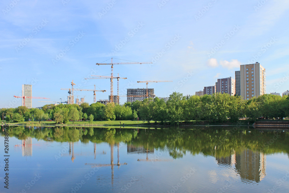 Fototapeta premium Construction of residential buildings with cranes against the blue sky in the early morning and reflected in the pond