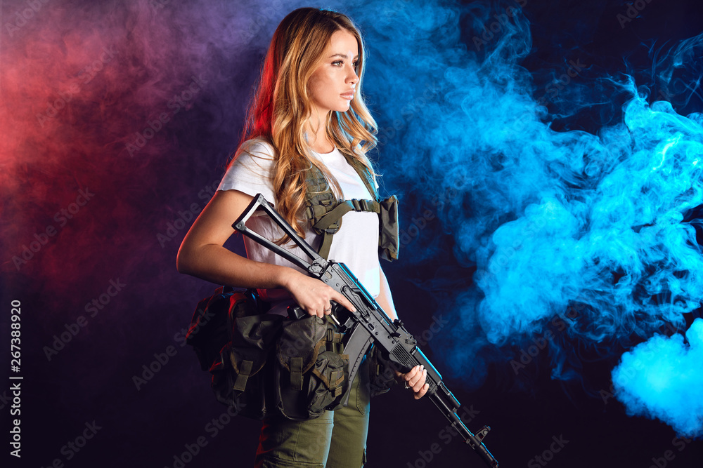 Woman soldier in military camouflage uniform protected with headphones, body armour, holding assault rifle standing against dark smoky background Woman soldier in military camouflage uniform protected with headphones, body armour, holding assault rifle standing against dark smoky background