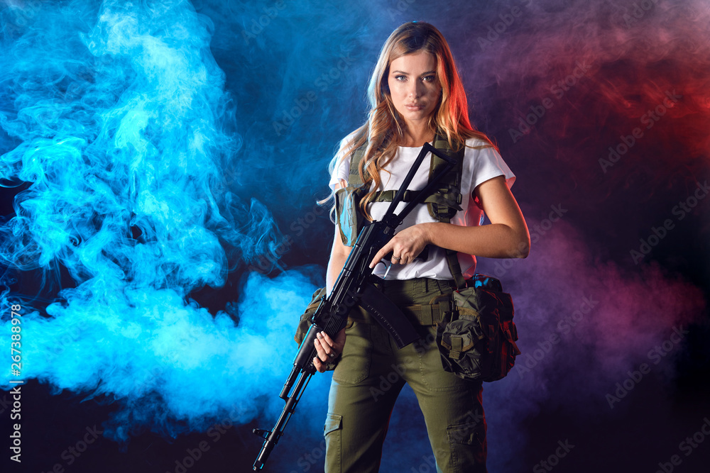 Young blonde female snipper in military outfit with assault rifle in studio on smoky dark background. Women in military service concept.