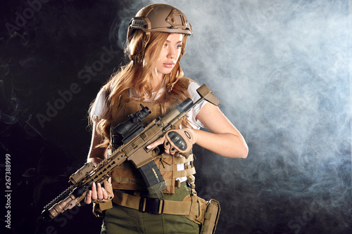 Beautiful caucasian female private military contractor with long blonde hair, dressed in tactical uniform and battle helmet, armed with riffle posing isolated in studio over black foggy background