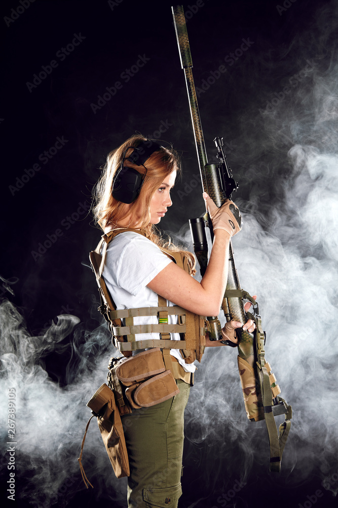 Skilled blonde female soldier with rifle in hands standing in military ...
