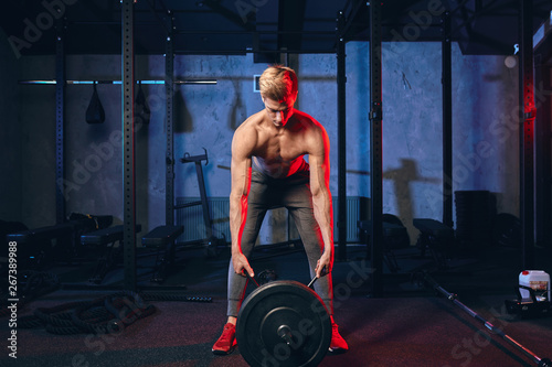 Caucasian muscular shirtless bodybuilder, flexing weight plates on iron bar over smoky dual color background. Weightlifting training.