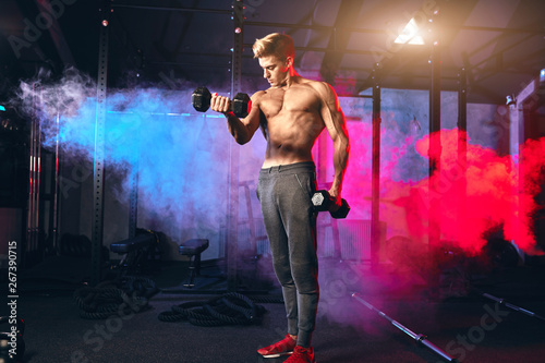 Shirtless sexy strong bodybuilder, athletic fitness man pumping up muscles with dumbbells in indoor gym workout against dark background.