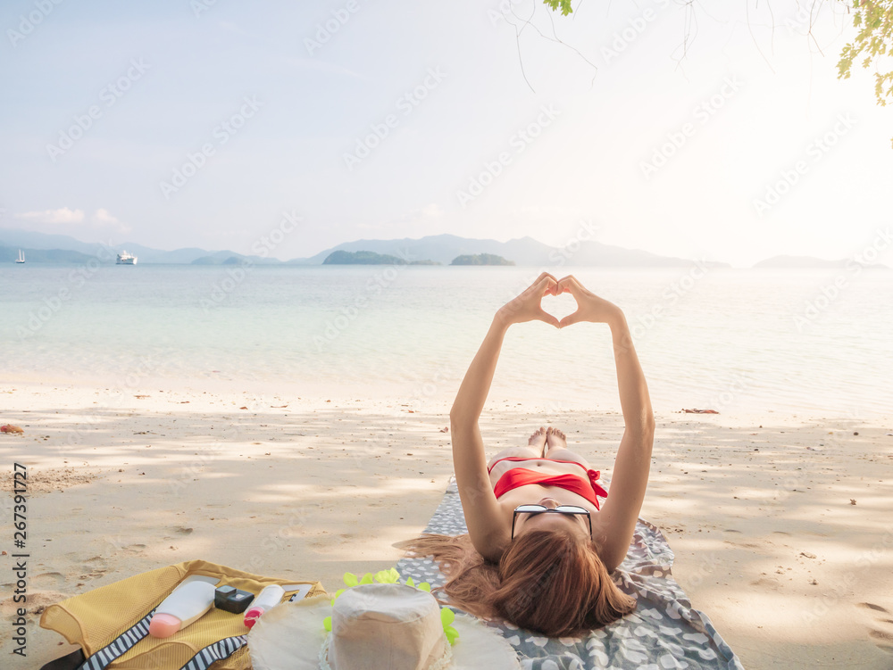 summer relax concept from young sexy woman in red bikini making hands in shape of love heart, lying on sandy tropical beach, for beauty skincare sun aging protection body care of tanned skin.