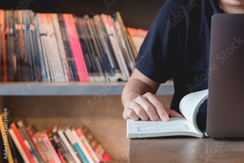 Young business man learning stock trading. Man sitting in library reading book finger pointing, studying book and connecting to internet using laptop making research.