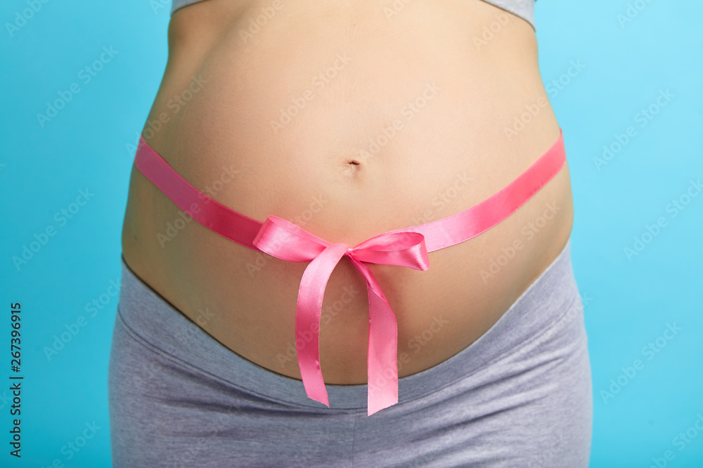 Close up of of pregnant woman tying up her tummy with pink ribbon on blue background Close up of of pregnant woman tying up her tummy with pink ribbon on blue background
