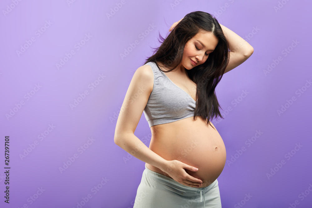 Pregnant woman in third trimester, dressed in comfotable home wear, posing against studio purple wall, looks happily at camera, being in anticipation of baby. Happy pregnancy and motherhood concept Pregnant woman in third trimester, dressed in comfotable home wear, posing against studio purple wall, looks happily at camera, being in anticipation of baby. Happy pregnancy and motherhood concept