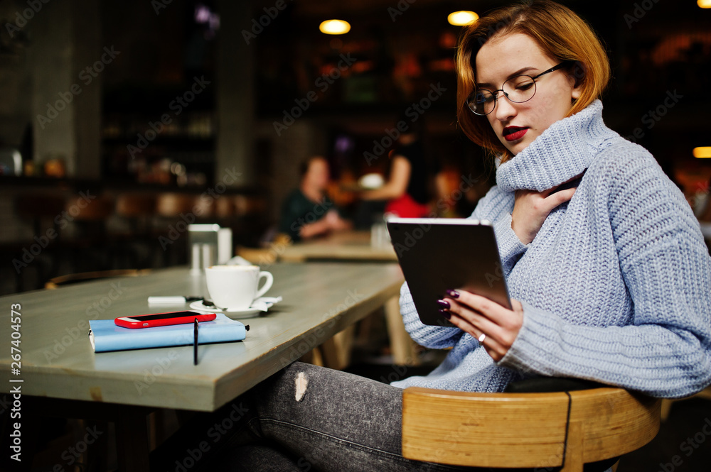 Cheerful young beautiful redhaired woman in glasses using her phone, touchpad and notebook while sitting at her working place on cafe with cup of coffee. Cheerful young beautiful redhaired woman in glasses using her phone, touchpad and notebook while sitting at her working place on cafe with cup of coffee.