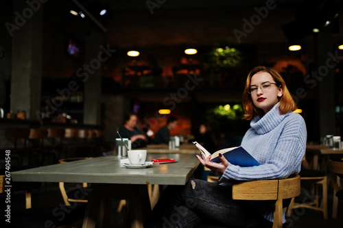 Cheerful young beautiful redhaired woman in glasses using her phone  touchpad and notebook while sitting at her working place on cafe with cup of coffee.