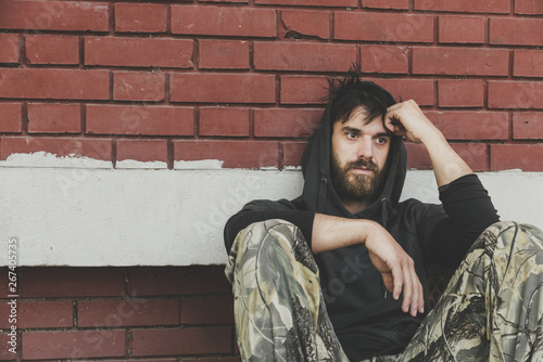 Homeless man, Homeless man drug and alcohol addict sitting alone and depressed on the street leaning against brick building wall feeling anxious and lonely, social documentary concept hard contrast 