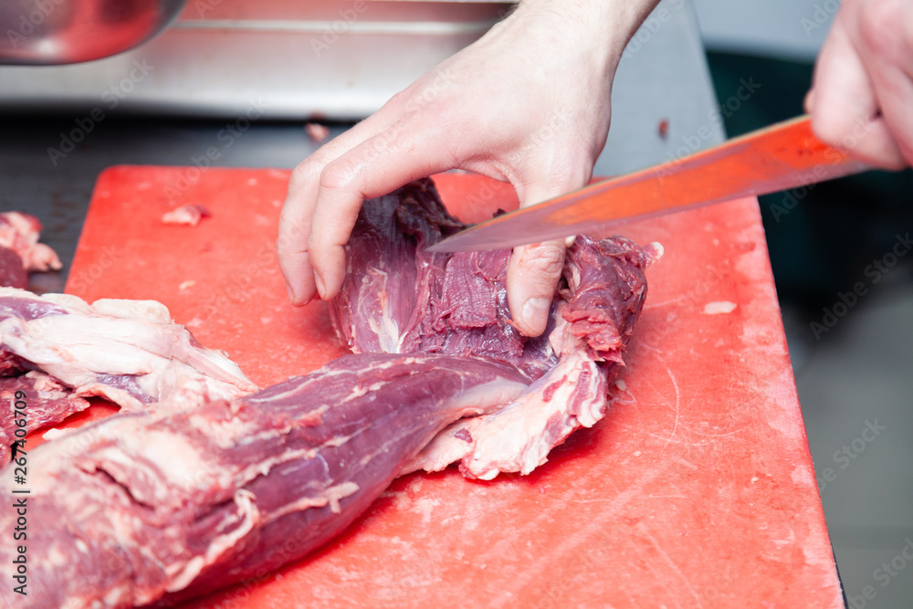 Closeup caucasian man chef butcher hands cutting piece of fillet raw pork tenderloin with knife, on plastic chopping board on professional table. Concept carve fresh meat grill on market