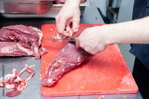 Closeup hands of man butcher chef cook cuts pork and beef meat fillet with professional knife in restaurant on red plastic board. Concept carve fresh meat barbeque grill on local market