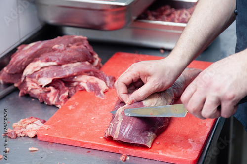 Closeup hands of man butcher chef cutting slices of fillet raw beef and pork meat off loin with knife in restaurant on red plastic board. Concept carve meat for barbeque grill on farmer market