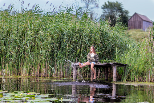 Happy little girl sitting on old wooden jetty surrounded by high green reeds splashing water with her feet at warm summer day. Summer vacation leisure time.