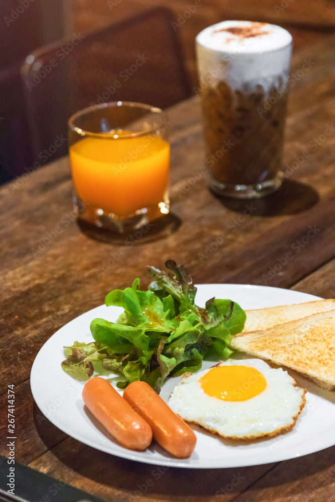 Fototapeta premium Delicious breakfast; fried egg, sausage, toast, salad with orange juice and iced coffee on wooden table.