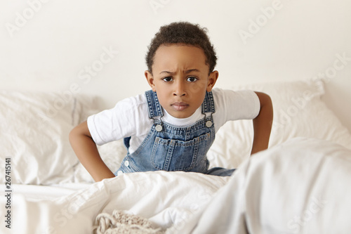 Human facial expressions and lifestyle concept. Indoor shot of grumpy black eyed African dark skinned sitting on cozy bed and frowning, being in bad mood, having angry look, not going to get up