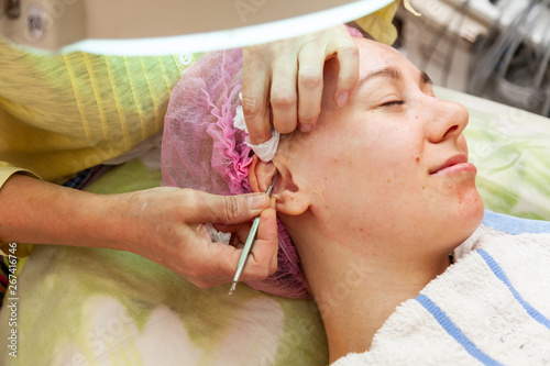A young girl is lying on a couch during cosmetic procedures with a mask on the face above which beautician woman squeezes body fat and pimples with special metal tool in the form of a blade in ear