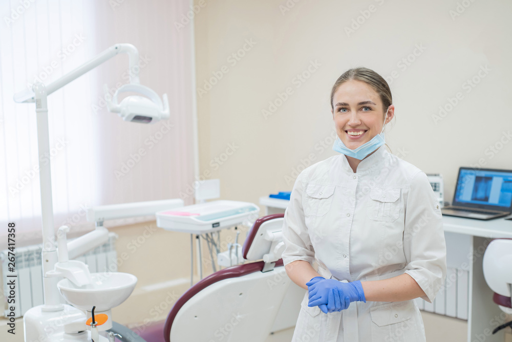 Attractive smiling female doctor in a white uniform at the workplace. A young beautiful female dentist in mask and gloves is standing in the office by the chair Attractive smiling female doctor in a white uniform at the workplace. A young beautiful female dentist in mask and gloves is standing in the office by the chair