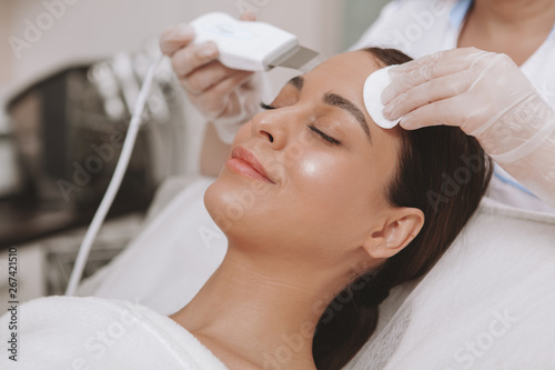 Young attractive woman smiling, receiving facial cleanse by professional cosmetologist. Beautician using hardware cosmetology equipment, working with female client. Relaxation, beauty concept photo