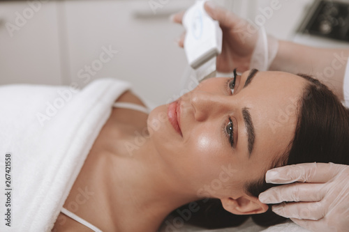 Beautiful happy woman smiling, getting ultrasound facial treatment by professional cosmetologist. Beautician cleaning face of a female patient with ultrasonic hardware equipment