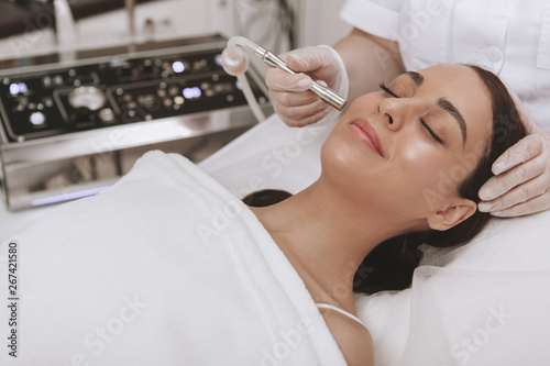 Professional cosmetologit performing microdermabrasion procedure on the face of a female client. Beautiful young woman getting face skin exfoliation treatment, copy space photo