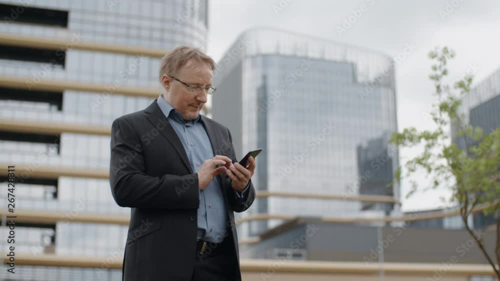 Funny man in suit and glasses texting on smartphone, adult blond manager or businessman standing on city street outside office talking cellphone and smiling, modern glass buildings business district Funny man in suit and glasses texting on smartphone, adult blond manager or businessman standing on city street outside office talking cellphone and smiling, modern glass buildings business district
