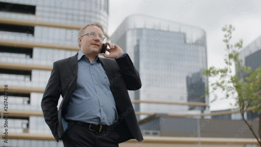 Funny plump man in suit and glasses talking by smartphone and smiling, adult blond manager or businessman standing on city street outside the office, modern glass buildings business district Funny plump man in suit and glasses talking by smartphone and smiling, adult blond manager or businessman standing on city street outside the office, modern glass buildings business district