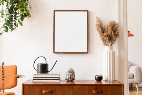 Stylish room of home interior with brown mock up frame with vintage accessories, plant, buddha,  orange chair and flowers in vase. Cozy home decor. Minimalistic concept. Retro composition of cupboard. © FollowTheFlow