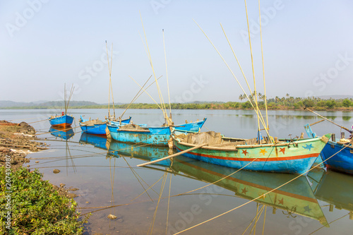 old blue large fishing boats with motors and fishing rods anchored off the coast against the backdrop of a river and green trees on the shore