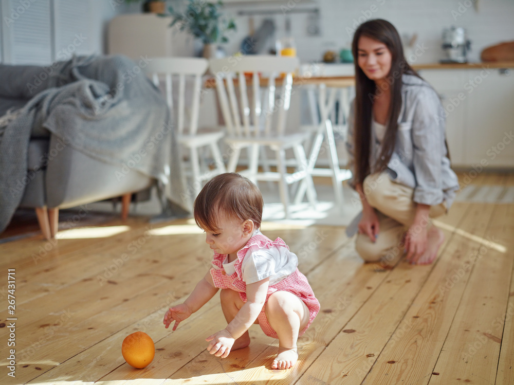 Fototapeta premium Young mother watching her cute baby daughter playing orange fruit on hardwood floor at home