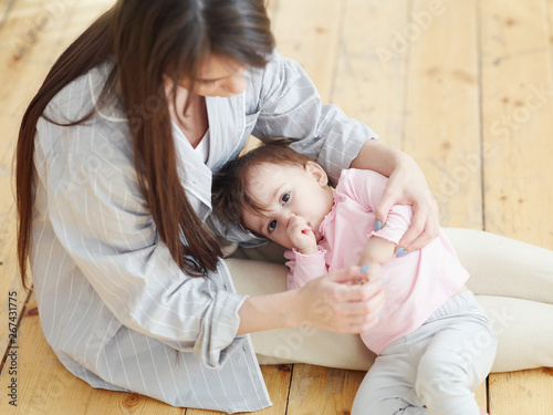 Cute baby girl looking at camera and sucking her thumb while lying on lap of young mother sitting on hardwood floor. Mom trying to break bad habit photo