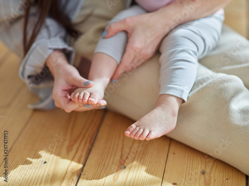 Hands of loving mother massaging cute foot of adorable baby daughter sitting on her lap