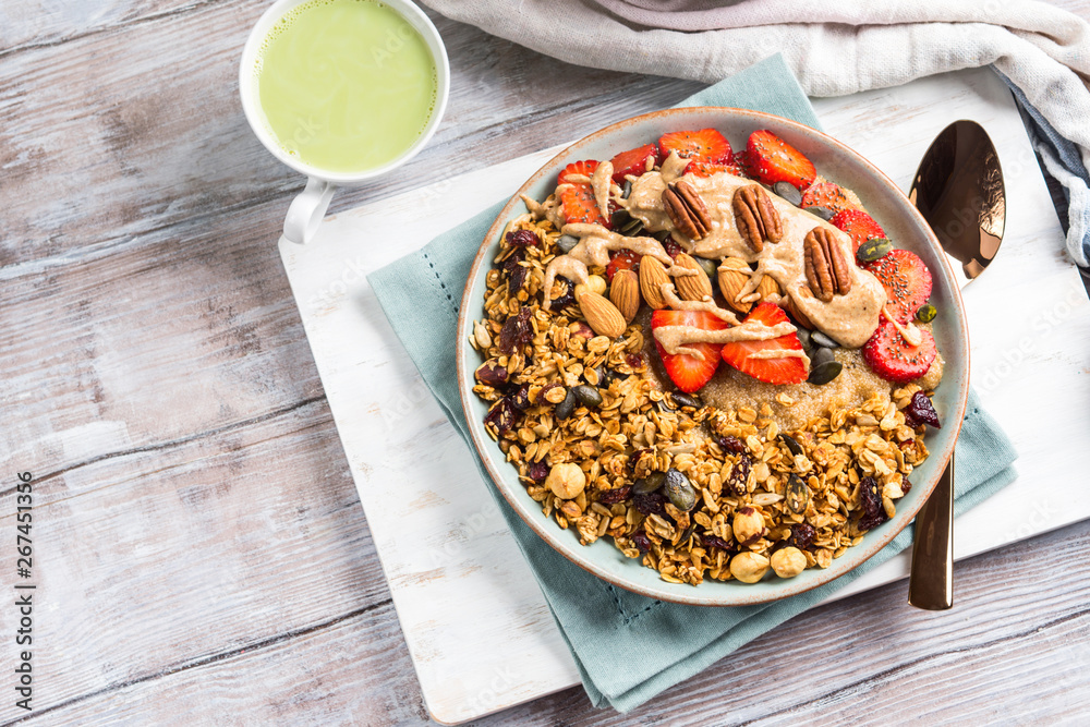 Fototapeta premium Delicious amaranth porridge topped with home made granola, strawberries and almond butter. Vegan plant based breakfast served with matcha latte on white wooden board. Healthy colorful food concept