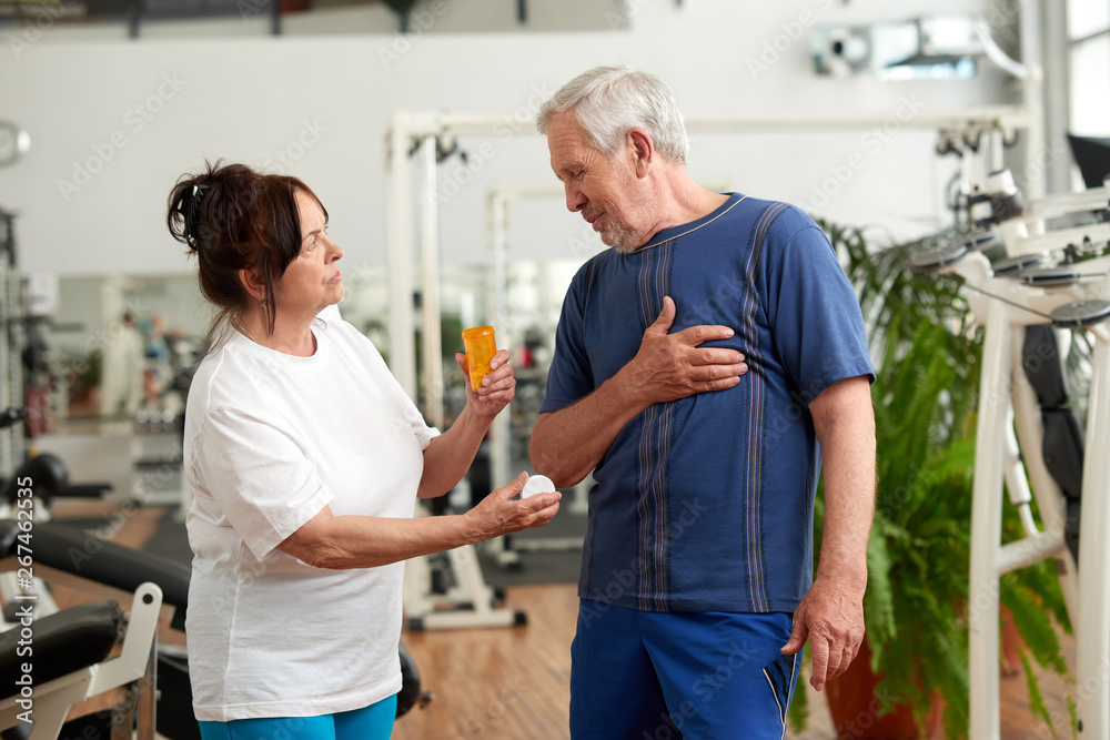 Senior people at fitness club. Elderly couple with opened bottle of pills at gym. People and health problem concept. Senior people at fitness club. Elderly couple with opened bottle of pills at gym. People and health problem concept.