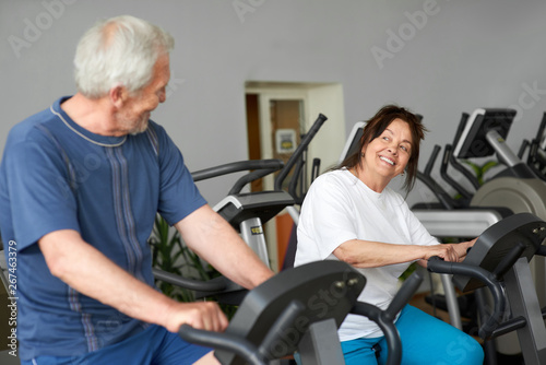 Beautiful couple of seniors working out at gym. Elderly man and woman training on machine at fitness club, focus on woman. Aged people doing cardio workout.