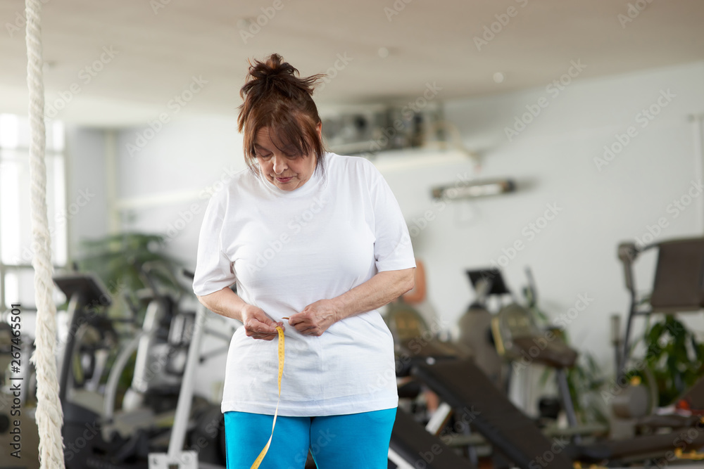 © DenisProduction.com - Senior woman with measuring tape at gym. Older caucasian woman measuring her waist by tape measure at fitness club. Sport for weight loss.
