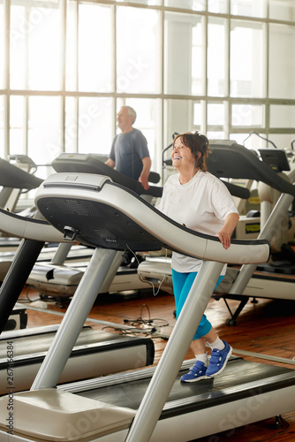 Smiling senior woman running on treadmill in gym. Cheerful aged woman in sportswear using treadmill machine at fitness center. People, fitness, active way of life. © DenisProduction.com