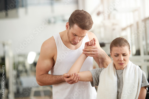 Man touching female painful elbow at gym. Young pretty woman with injured hand getting first aid from fitness trainer in gym. Sports exercising injury.