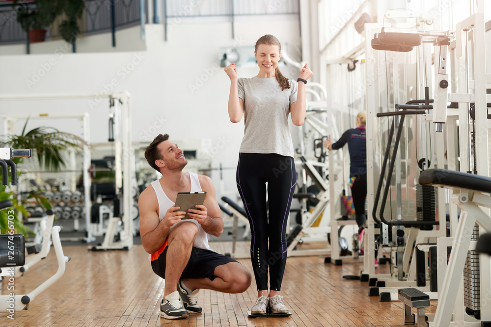 Excited young woman on weight scale at gym. Result of work with personal trainer. Slimming and dieting. Healthy lifestyle concept. Excited young woman on weight scale at gym. Result of work with personal trainer. Slimming and dieting. Healthy lifestyle concept.
